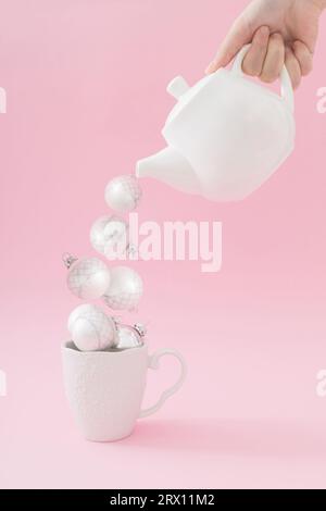 Composition créative avec main tenant la théière et versant des boules de Noël dans une tasse à thé sur fond rose. Concept minimal de Noël ou de nouvel an. Banque D'Images