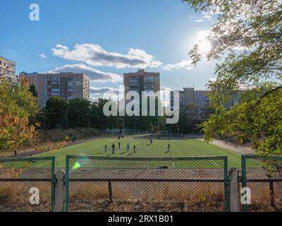 Les enfants jouent au soccer sur un terrain de sport vert, une aire d'entraînement avec équipement d'exercice en métal entourée de gratte-ciel dans une communauté résidentielle. Banque D'Images