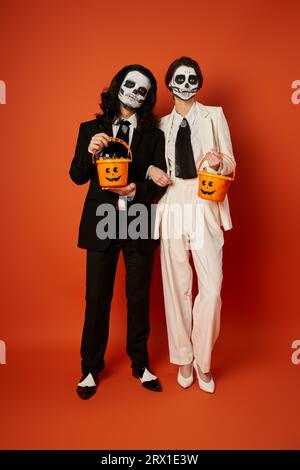 Toute la longueur d'un couple élégant en maquillage catrina tenant des seaux de bonbons sur le rouge, Day of Dead fest Banque D'Images