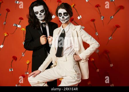 Couple en maquillage catrina et tenue élégante posant sur fond rouge avec des fleurs, Day of Dead Banque D'Images