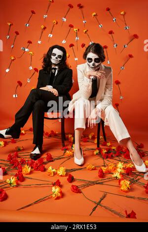 pleine longueur de couple élégant dans le maquillage de crâne et costumes assis sur des chaises dans le studio rouge avec des fleurs Banque D'Images