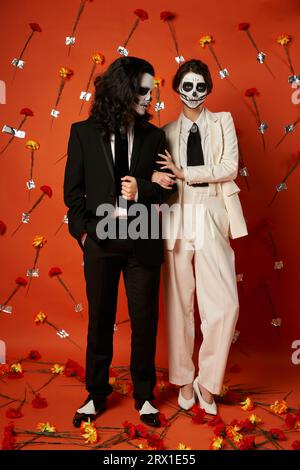 toute la longueur du couple en maquillage catrina et tenue de fête posant sur fond rouge avec œillets Banque D'Images