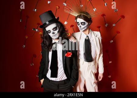 couple élégant en dia de los muertos crâne maquillage regardant la caméra sur fond rouge avec des fleurs Banque D'Images