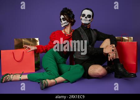 couple élégant en dia de los muertos crâne maquillage assis près des sacs à provisions sur bleu, pleine longueur Banque D'Images