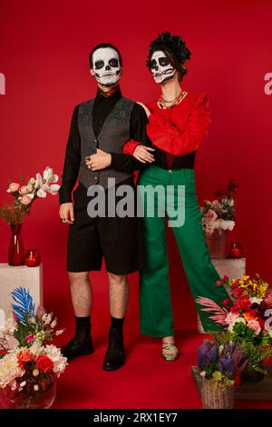 Pleine longueur de couple dans le maquillage de crâne près traditionnel Day of Dead ofrenda avec des fleurs sur rouge Banque D'Images