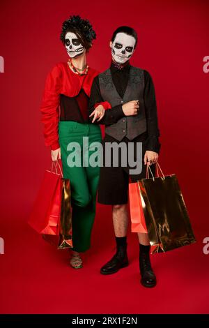 pleine longueur de couple tendance en maquillage de crâne de sucre tenant des sacs à provisions sur rouge, dia de los muertos Banque D'Images