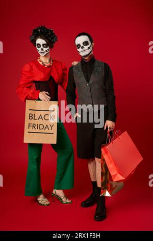 pleine longueur de couple à la mode dans le maquillage de crâne de sucre tenant des sacs à provisions le rouge, vendredi noir Banque D'Images