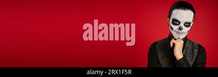 portrait de l'homme en dia de los muertos sucre crâne maquillage tenant la main près du menton sur rouge, bannière Banque D'Images