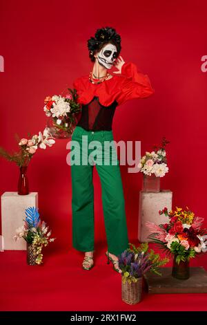 femme dans le maquillage de crâne de sucre près de l'autel traditionnel dia de los muertos avec des fleurs colorées sur le rouge Banque D'Images