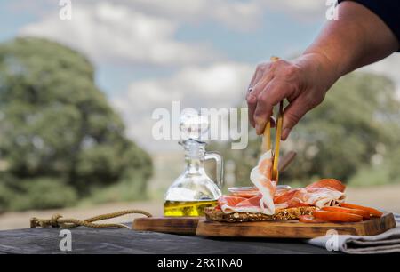 Femme mettant des tranches de jambon ibérique nourri aux glands sur une tranche de pain avec de la tomate et de l'huile d'olive sur une table en bois à la campagne, typiquement espagnole Banque D'Images