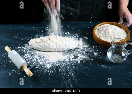 Femme versant de la farine sur une pâte fraîche pour faire du pain maison sur une table en bois avec de la farine, de l'eau et un rouleau à pâtisserie Banque D'Images