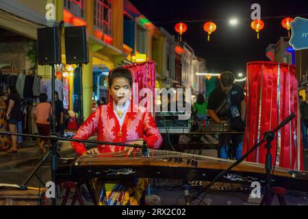 Phuket, Thaïlande - 21 février 2018 : jeune femme en robe rouge jouant de la cithare pincée dans la rue nocturne de la ville, nouvel an chinois. Femme de beauté cueillir woo Banque D'Images