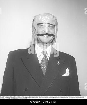 Dans les années 1950 C'est un casque étrange que l'homme en costume porte sur sa tête. Le modèle est en plastique avec une matière plastique transparente devant les yeux. On peut spéculer sur la façon dont il a été utilisé, mais une hypothèse est que c'est une sorte de bonnet de douche qui protège les cheveux de l'eau. La photo a été prise lors d'un salon des consommateurs où le produit a été présenté au public pour la première fois. Le plastique était le nouveau matériau des années 50 qui était utilisé dans tout, des ustensiles ménagers à la décoration intérieure. Suède 1951. Kristoffersson réf. BD39-9 Banque D'Images