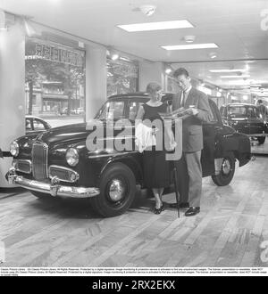 Dans les années 1950 Un jeune couple visite un concessionnaire automobile et le concessionnaire qui vend la marque anglaise Humber. Le couple étudie un dossier sur la voiture dans la pièce où la voiture est garée et semble satisfait. Ils lisent un dossier sur le voyage emblématique avec la voiture entre Londres et Cape Town. En 1952, George Hinchcliffe, Arthur Longman et Robbie Walshaw partirent de Londres pour se rendre au Cap dans un Humber Supersnipe dans le temps record de 13 jours 9 heures 6 minutes. Suède 1954. Kristoffersson réf BP39-5 Banque D'Images