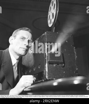 Homme à un projecteur de film dans les années 1940 Un homme devant un projecteur de cinéma qui tourne. Il a les yeux sur l'écran où le film est projeté. Le film était généralement montré sur un écran blanc. Vous pouvez voir la bobine de film en haut et la bande de film introduite dans le projecteur à une vitesse précise. Suède 1943. Kristoffersson réf. B20-3 Banque D'Images