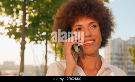 Close up souriant afro-américaine heureuse femme d'affaires ethnique marchant dans le soleil soleil ville à distance parler parler téléphone portable sourire fin de fin Banque D'Images