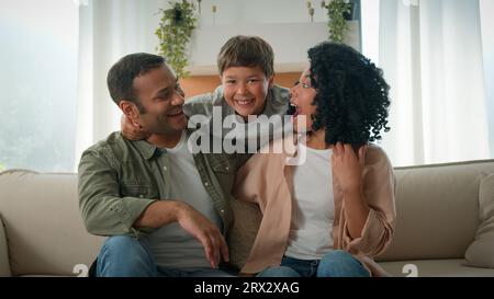 Affectueux parents adulte Afro-américain mère et père embrassent câlin sur le canapé drôle petit garçon adopté fils enfant enfant sautant derrière maman câlin Banque D'Images