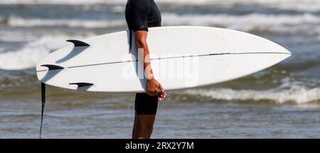 Vue latérale d'un homme tenant une planche de surf blanche sur la plage avec l'océan Atlantique en arrière-plan à Gilgo Beach long Island New York. Banque D'Images