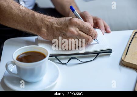 Les mains du vieil homme écrivent des notes tout en buvant du café dans le salon Banque D'Images