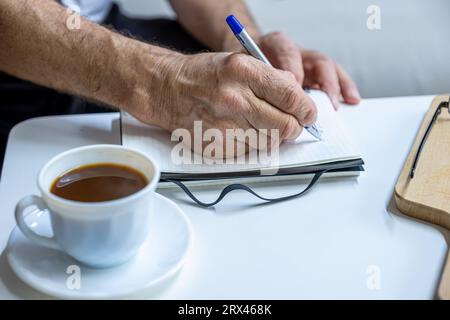 Les mains du vieil homme écrivent des notes tout en buvant du café dans le salon Banque D'Images