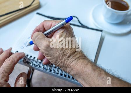 Les mains du vieil homme écrivent des notes tout en buvant du café dans le salon Banque D'Images