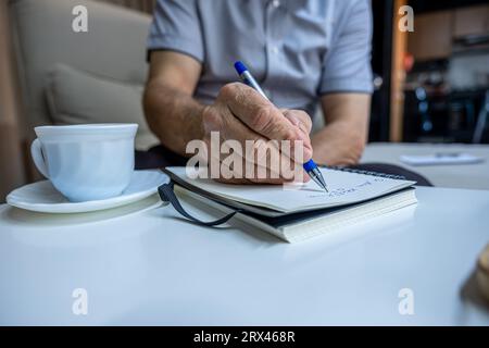 Les mains du vieil homme écrivent des notes tout en buvant du café dans le salon Banque D'Images