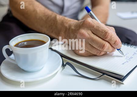 Les mains du vieil homme écrivent des notes tout en buvant du café dans le salon Banque D'Images