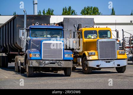 Jaune et bleu vieux style cabine de jour gros semi-camions de forage tracteurs avec des remorques basculantes à benne basculante debout dans la rangée sur les emplacements marqués du parking de l'entrepôt Wai Banque D'Images