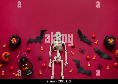 Composition avec des cors de bonbons savoureux, squelette, lanterne et décor d'Halloween sur fond rouge Banque D'Images