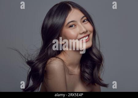 Jolie femme asiatique aux cheveux longs et brillants ondulés sur un gris isolé. Banque D'Images