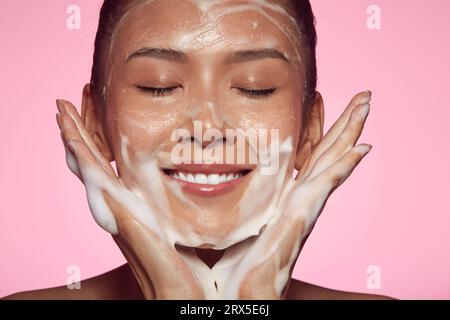 Jeune femme asiatique utilisant la mousse nettoyante de peau de soin quotidien et regardant ravie debout sur fond isolé rose. Banque D'Images