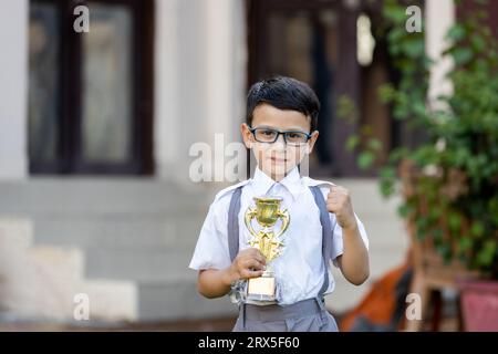 Heureux écolier indien portant l'uniforme scolaire tenant le trophée de victoire en main à l'extérieur de l'école. Concept d'éducation. Banque D'Images