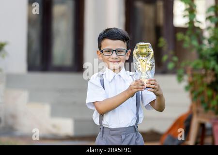 Heureux écolier indien portant l'uniforme scolaire tenant le trophée de victoire en main à l'extérieur de l'école. Concept d'éducation. Banque D'Images