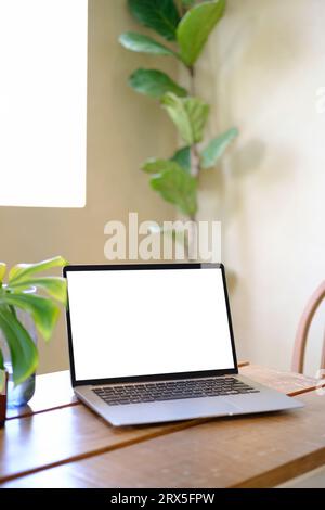 Ordinateur portable avec écran blanc vide et feuilles de palmier tropical monstera dans un vase en verre sur une table en bois Banque D'Images