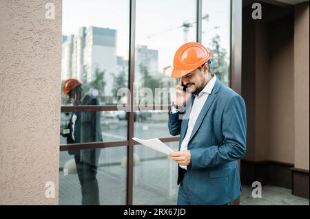 Ingénieur souriant parlant sur le téléphone intelligent devant le bâtiment Banque D'Images