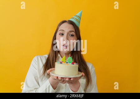 Femme portant le chapeau de fête tenant le gâteau d'anniversaire sur fond jaune Banque D'Images