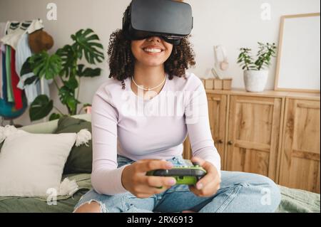 Fille heureuse portant des simulateurs de réalité virtuelle tenant le contrôleur de jeu sur le lit à la maison Banque D'Images