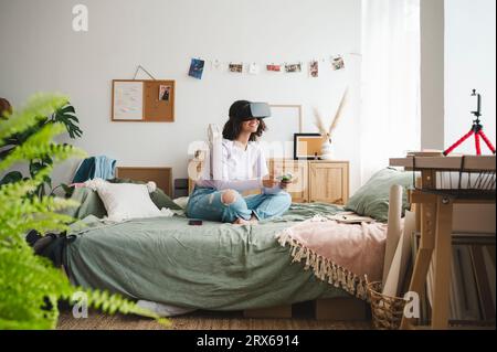 Fille souriante portant des simulateurs de réalité virtuelle tenant le contrôleur de jeu sur le lit à la maison Banque D'Images