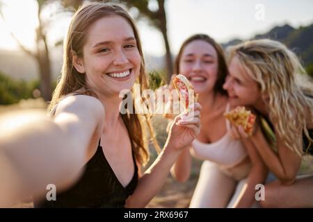 Femme heureuse ayant pizza et prenant selfie avec des amis dans le parc Banque D'Images