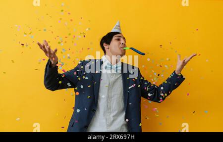 Jeune homme soufflant de la corne de fête et jetant des confettis en studio Banque D'Images