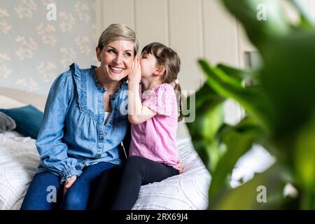 Fille chuchotant dans l'oreille souriante de la mère dans la chambre Banque D'Images