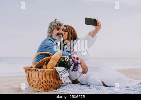 Heureuse femme senior embrassant l'homme et prenant selfie par téléphone intelligent à la plage Banque D'Images