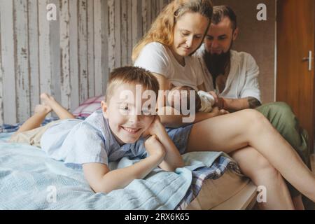 Fils souriant couché à côté des parents avec la fille nouveau-née sur le lit à la maison Banque D'Images