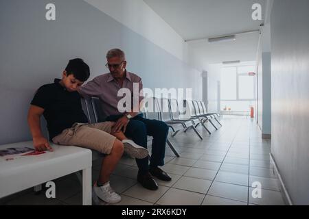 Enfant avec son grand-père attendant leur tour dans un hôpital tout en étant assis sur un banc dans le hall et ayant une conversation. Grand-père réconfortant ne Banque D'Images