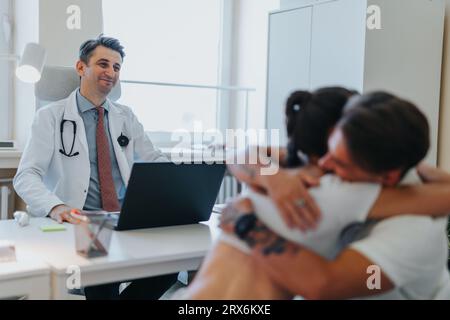 Heureux, médecin souriant informe le jeune couple de leur grossesse. Couple s'embrassant après une information positive de grossesse par le médecin. Banque D'Images
