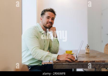 Jeune homme d'affaires souriant assis avec ordinateur portable dans le café Banque D'Images