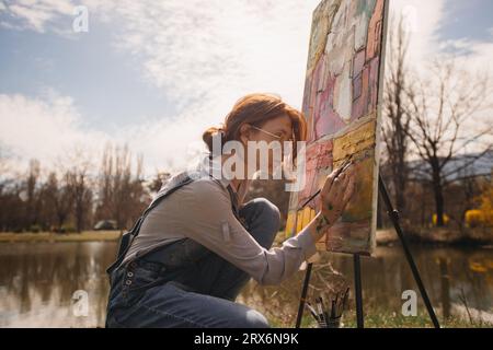 Belle fille avec les cheveux rouges et les lunettes peignant une image colorée sur une grande toile près du lac Banque D'Images