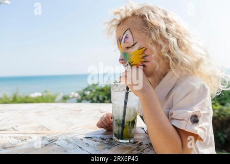 Fille blonde avec la peinture de visage buvant mojito à table Banque D'Images