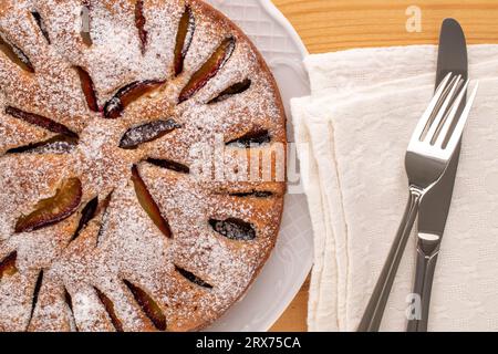 Gâteau aux prunes maison sucrée avec plat en céramique blanche, couteau et fourchette sur table en bois, macro, vue de dessus. Banque D'Images