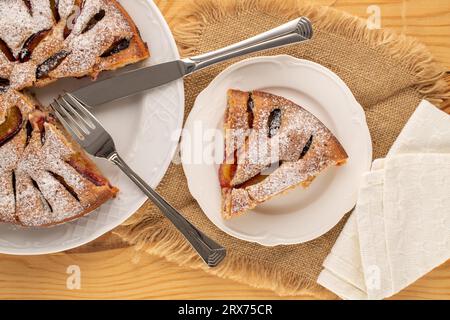 Gâteau aux prunes maison sucrée avec plat en céramique blanche, couteau et fourchette sur table en bois, macro, vue de dessus. Banque D'Images